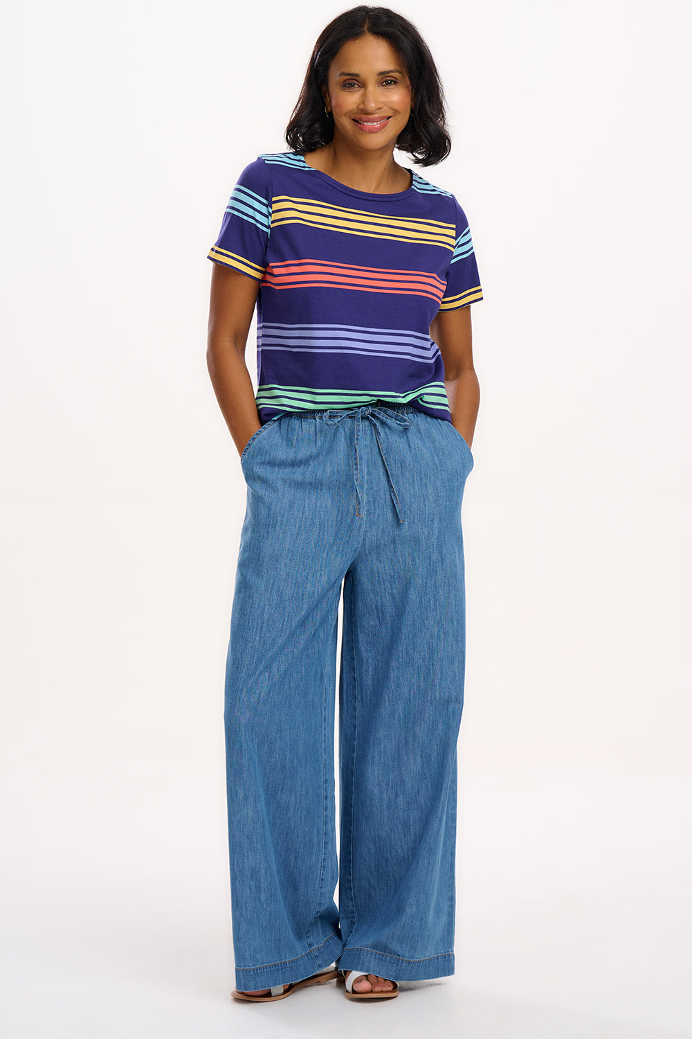 A woman wearing a navy t-shirt with wide muted stripes in mustard, coral, mint green, and baby blue over a navy base, paired with blue jeans.