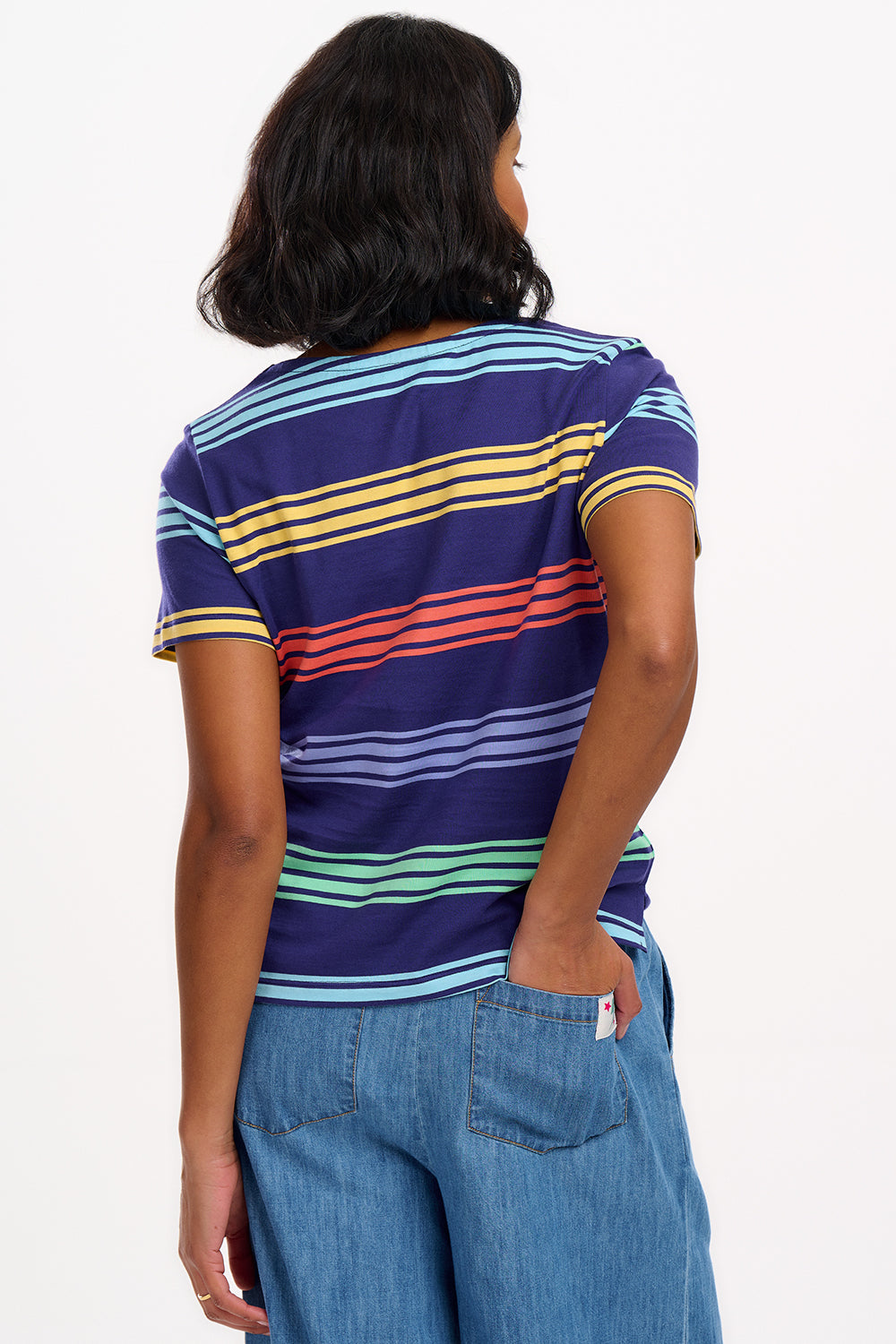 A woman wearing a navy t-shirt with wide muted stripes in mustard, coral, mint green, and baby blue over a navy base, paired with blue jeans.