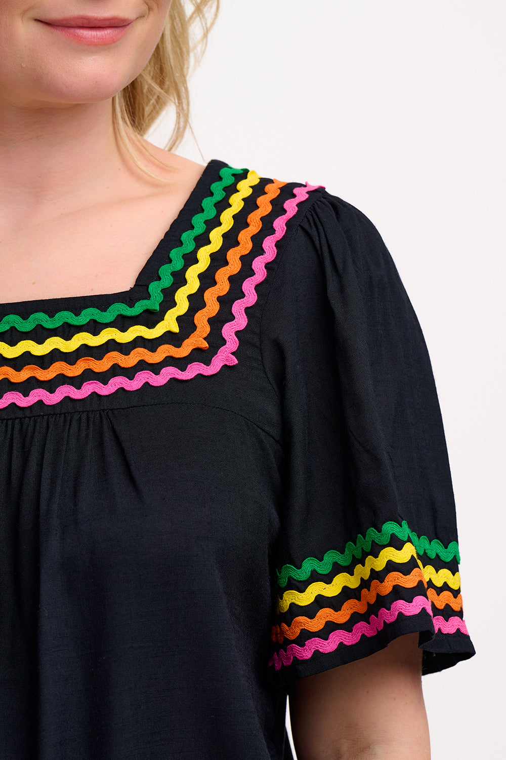 A woman wearing a black top with rainbow-colored ric rac detailing around the square neckline.