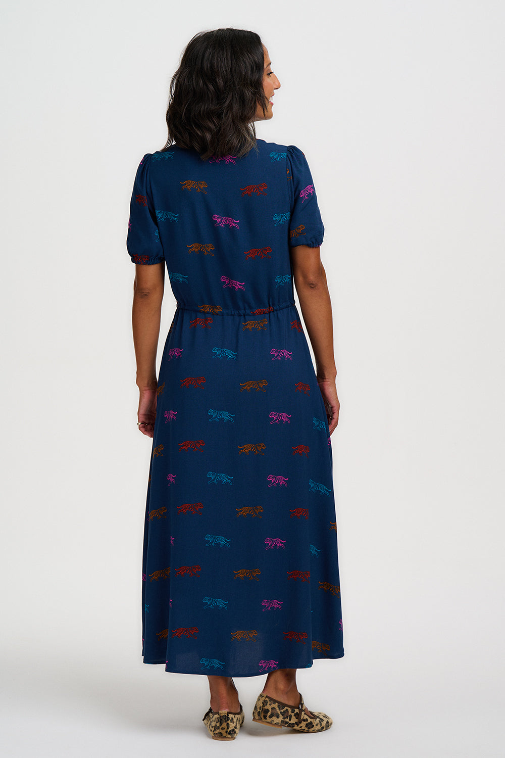 A woman viewed from the back wearing a navy midi dress with short puff sleeves and an allover multicolor dragonfly print, paired with leopard-print flats, against a light gray studio background.