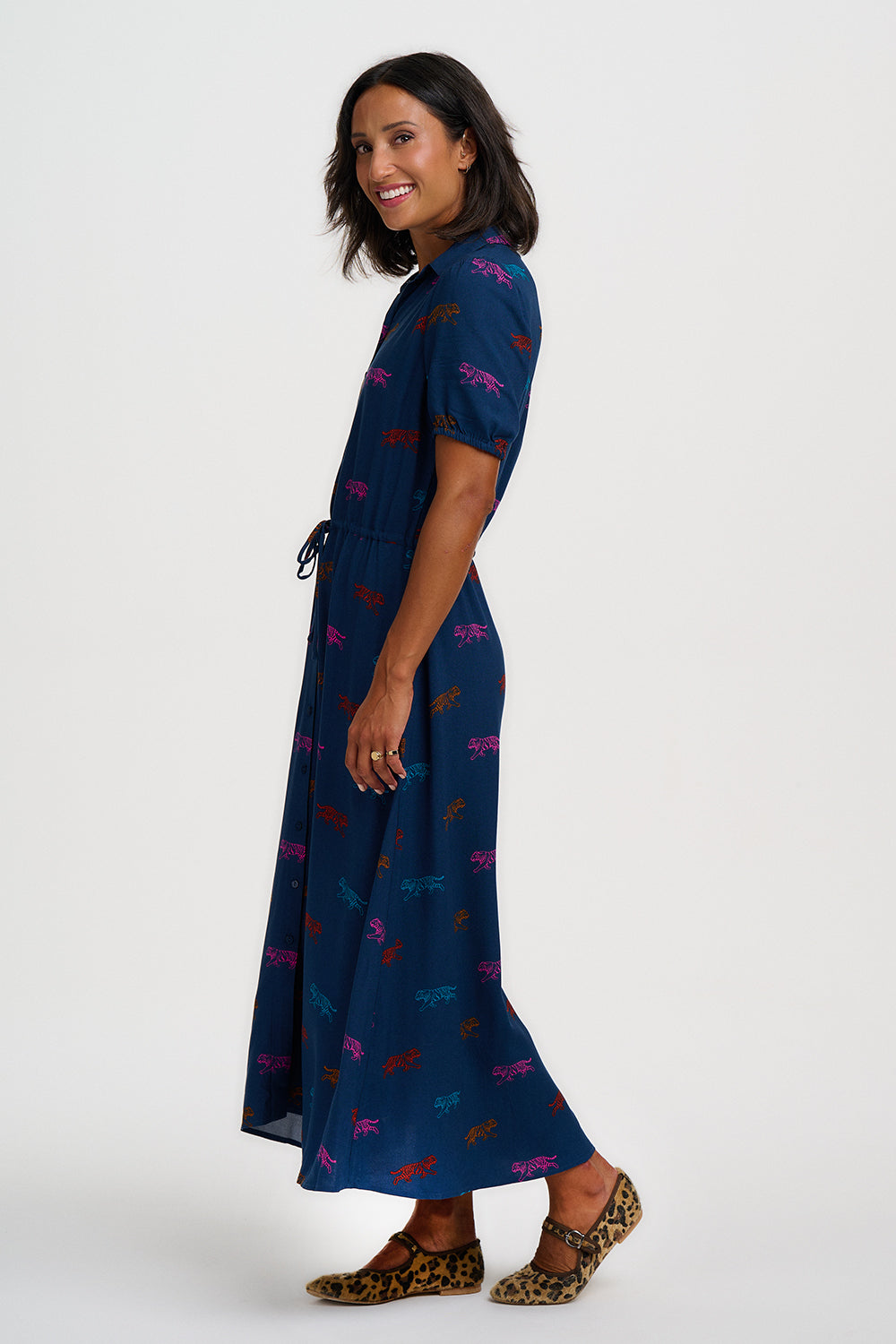 A woman modeling a navy blue maxi dress with a colorful abstract print, shown from a side-back angle against a light gray studio background.