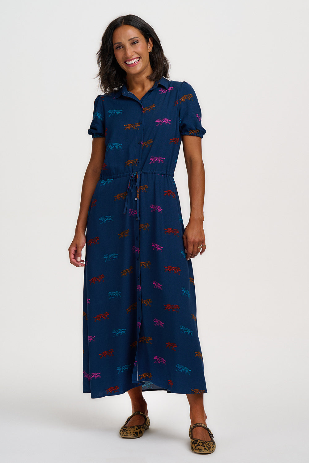A woman modelling a navy blue midi dress with short sleeves, a collared neckline, and an allover pattern of small colourful tigers. She is wearing leopard-print flat shoes against a plain light grey studio background.
