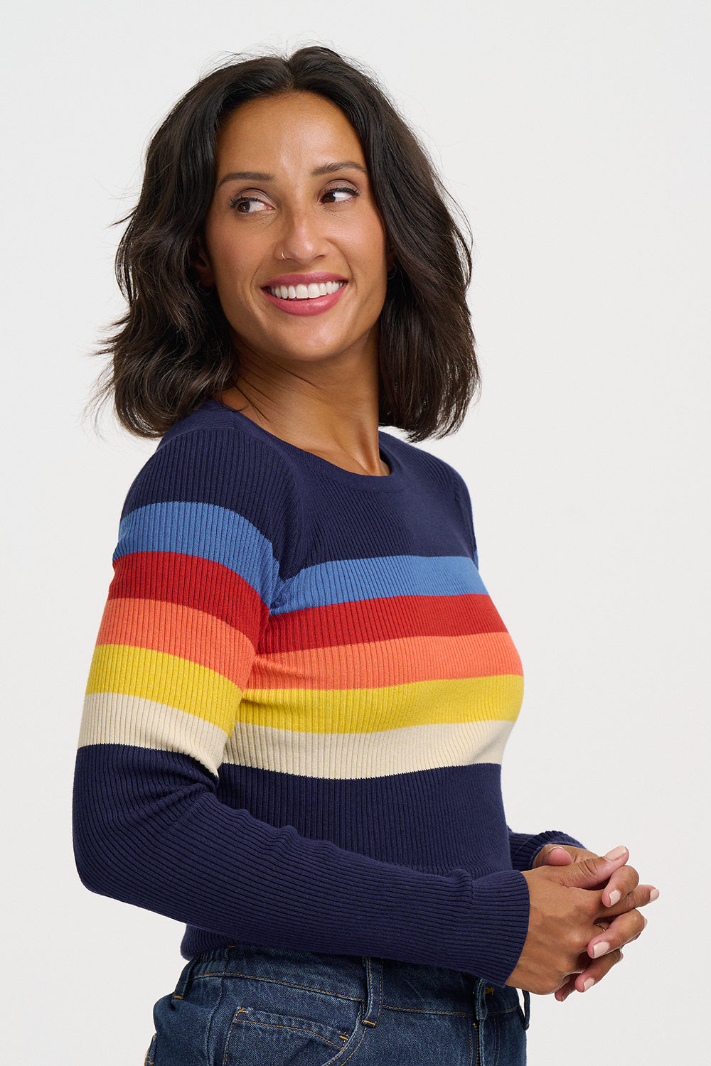 A woman wearing a navy blue ribbed knit sweater with horizontal stripes in light blue, red, orange, yellow, and cream, paired with blue jeans, against a light gray studio background.
