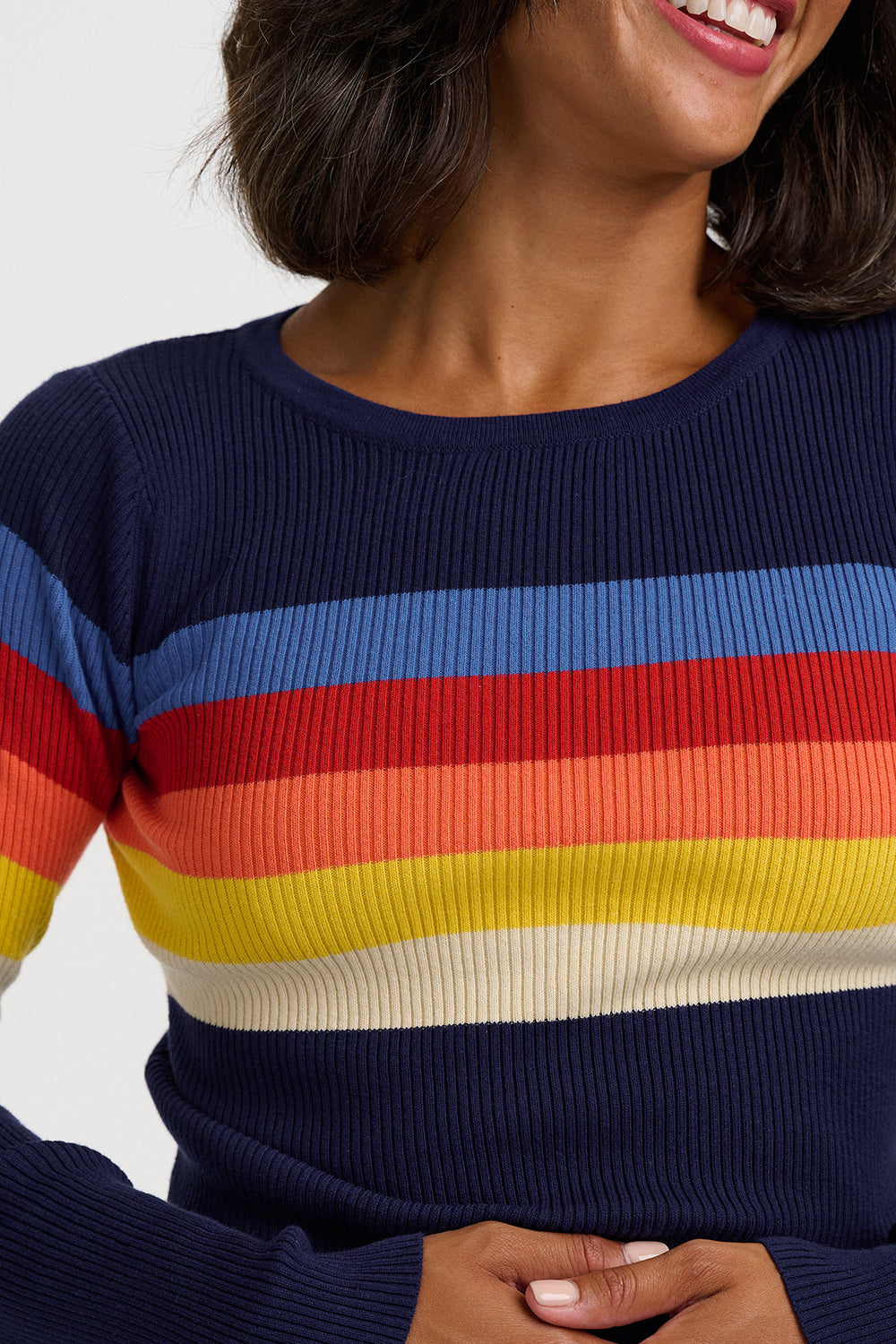 A close-up of a woman wearing a ribbed long-sleeve sweater with horizontal rainbow stripes in blue, red, orange, yellow, and cream, set against a plain light gray background.