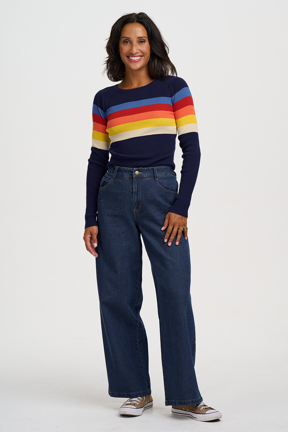 A woman modeling a navy blue long-sleeve knit sweater with horizontal stripes in red, orange, yellow, and white across the chest and sleeves, paired with dark blue wide-leg jeans and brown sneakers, against a light gray studio background.