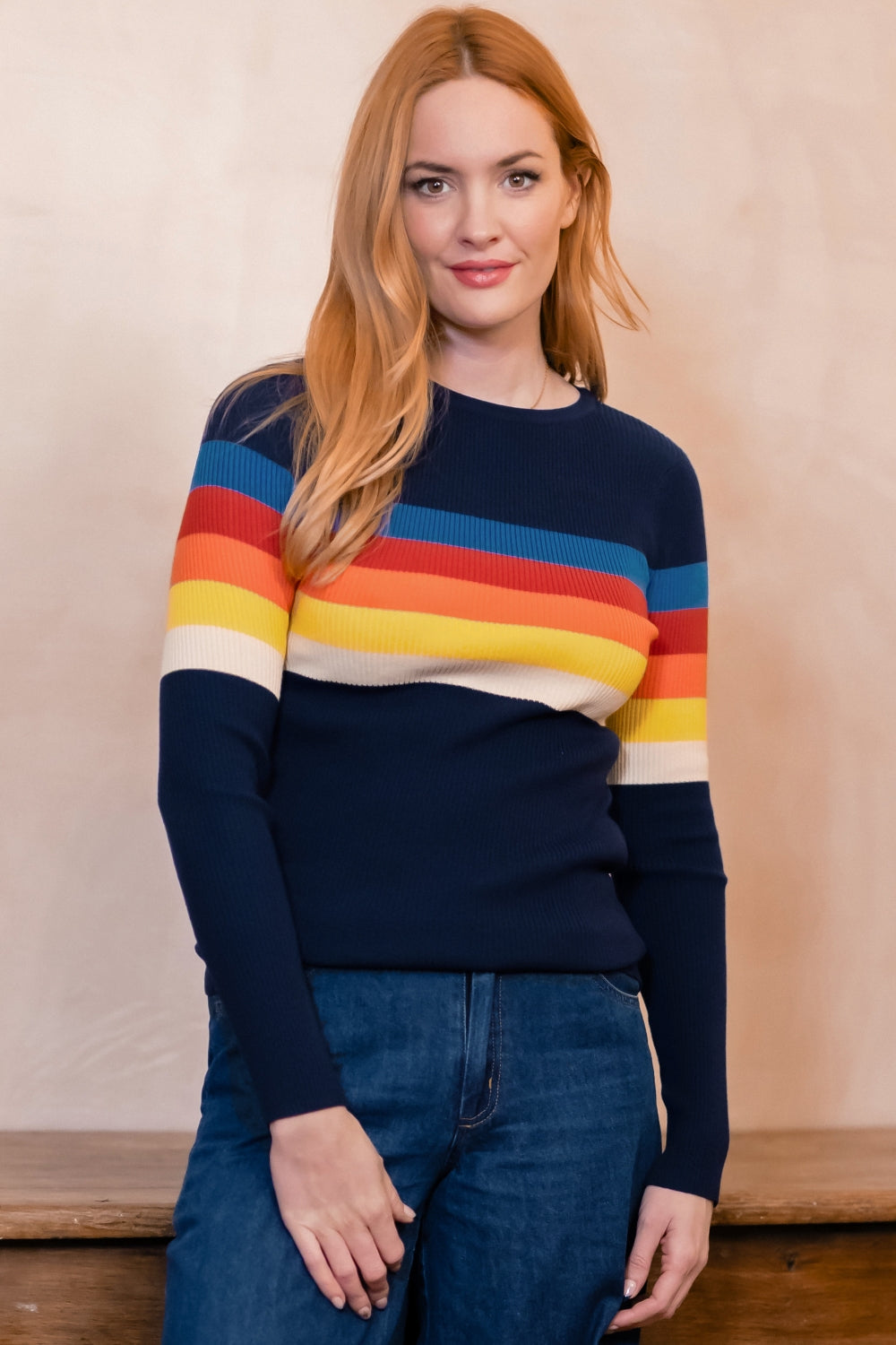 A woman with red hair wearing a navy blue crewneck sweater with horizontal rainbow stripes across the chest and sleeves, paired with blue jeans, standing against a warm beige textured wall.