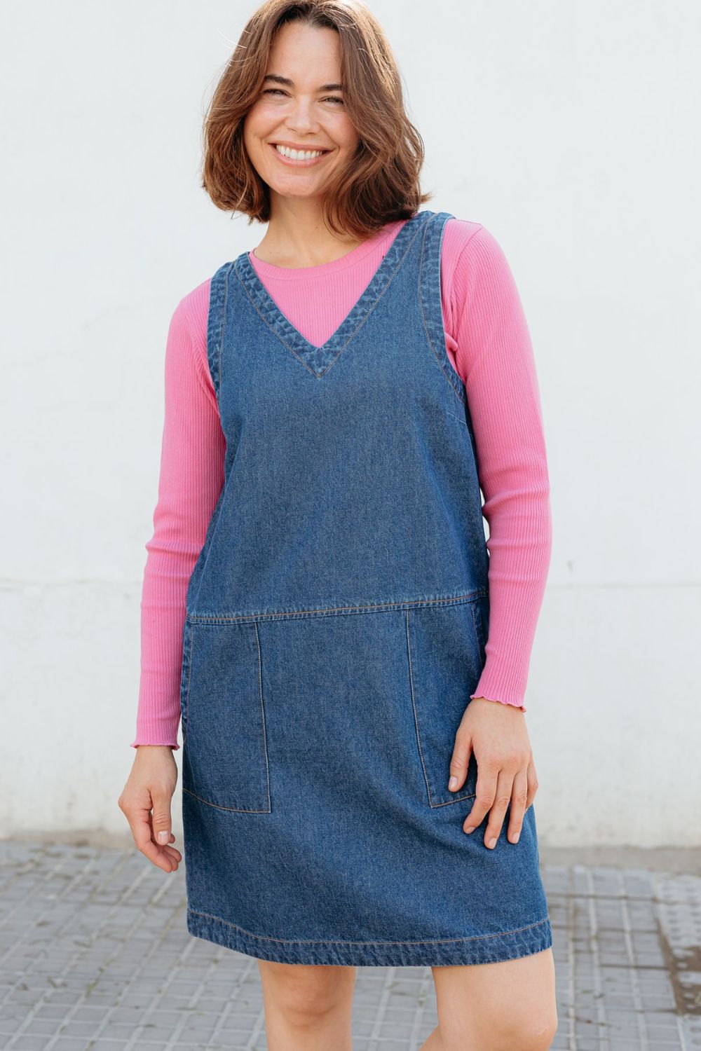 Woman wearing a blue denim dress over a pink long-sleeve shirt against a white wall.