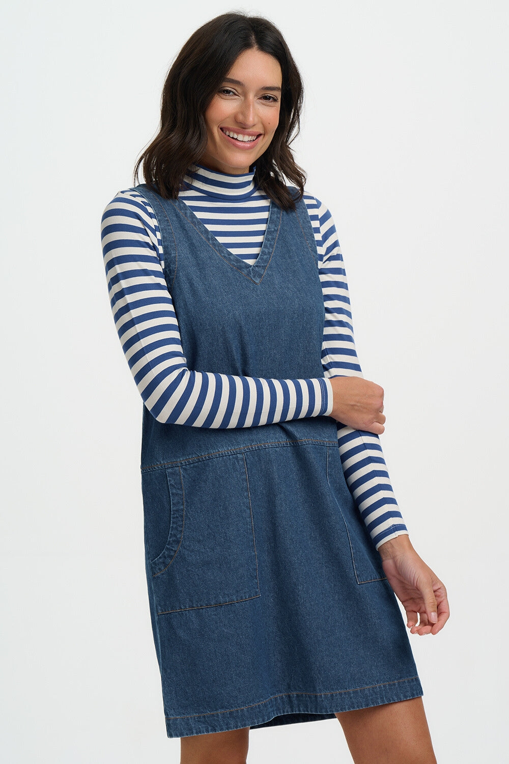 Woman wearing a blue denim pinafore dress over a striped long-sleeve shirt on a white background