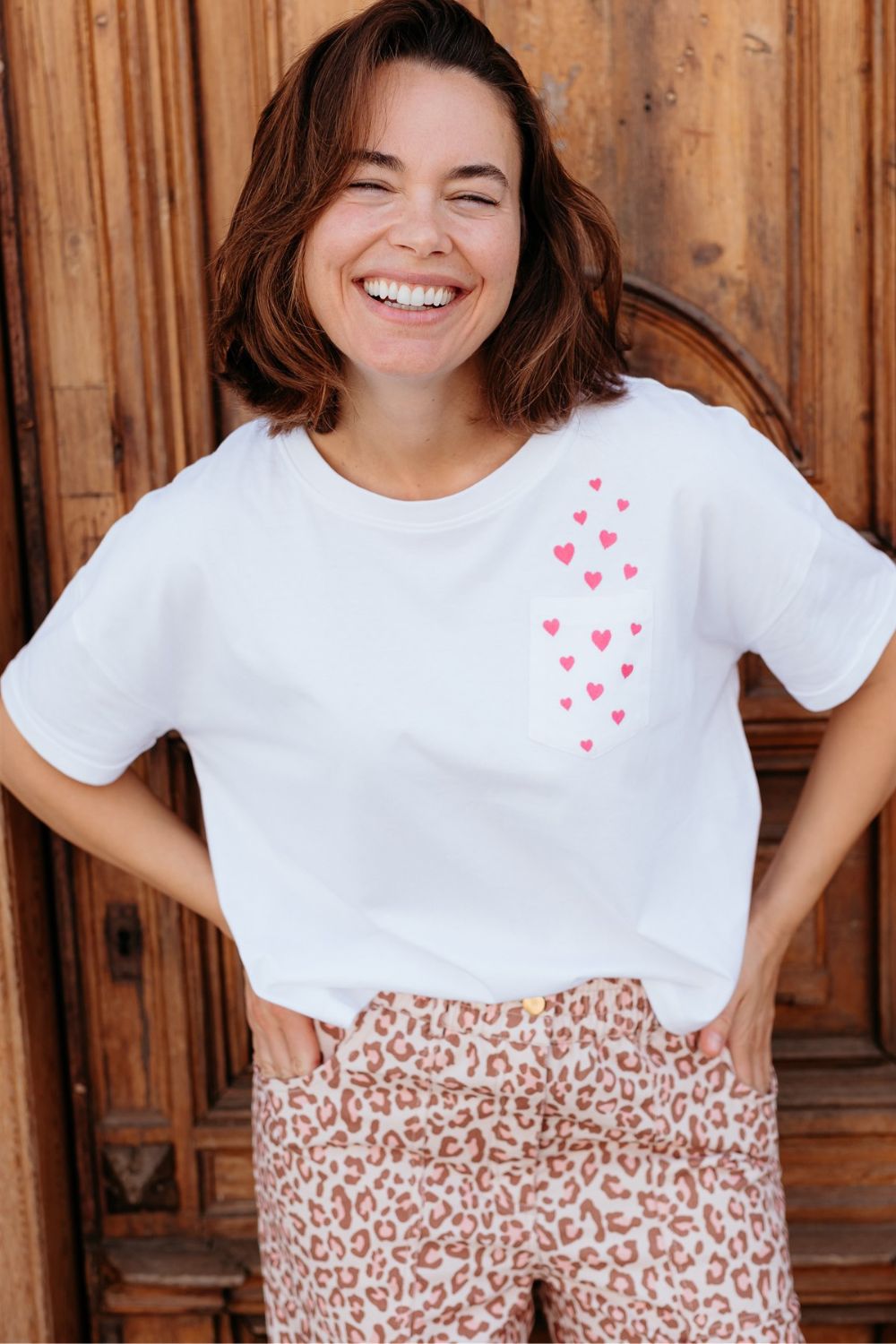 Woman wearing a white t-shirt with red heart pattern and leopard print pants standing against a wooden background