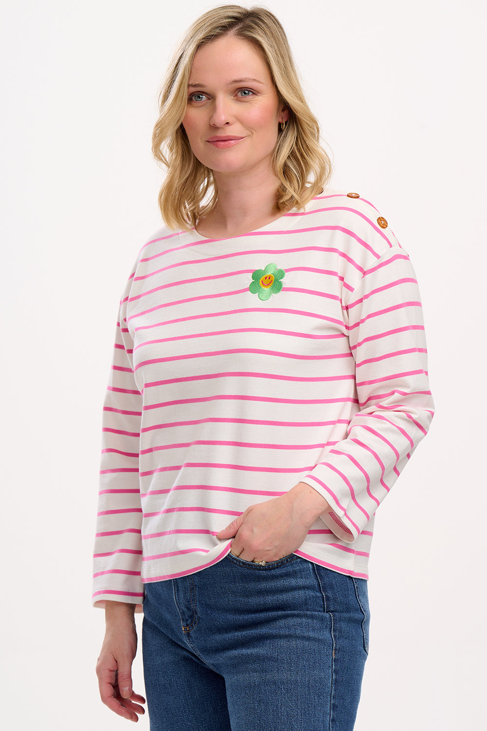 A woman wearing an off-white and pink striped Breton top with a smiley flower embroidery on the chest, paired with jeans.