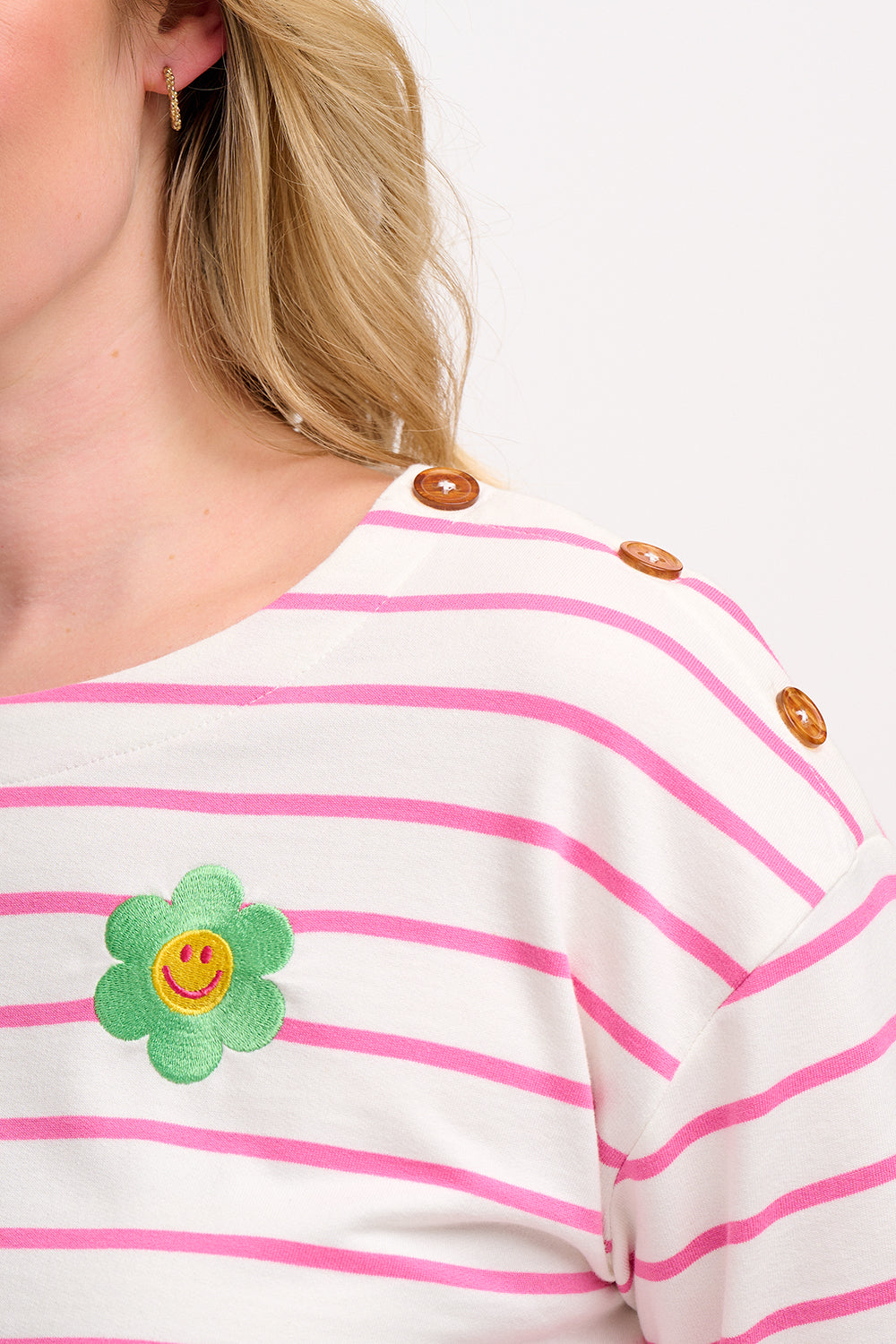 A woman wearing an off-white and pink striped Breton top with a smiley flower embroidery on the chest, paired with jeans.