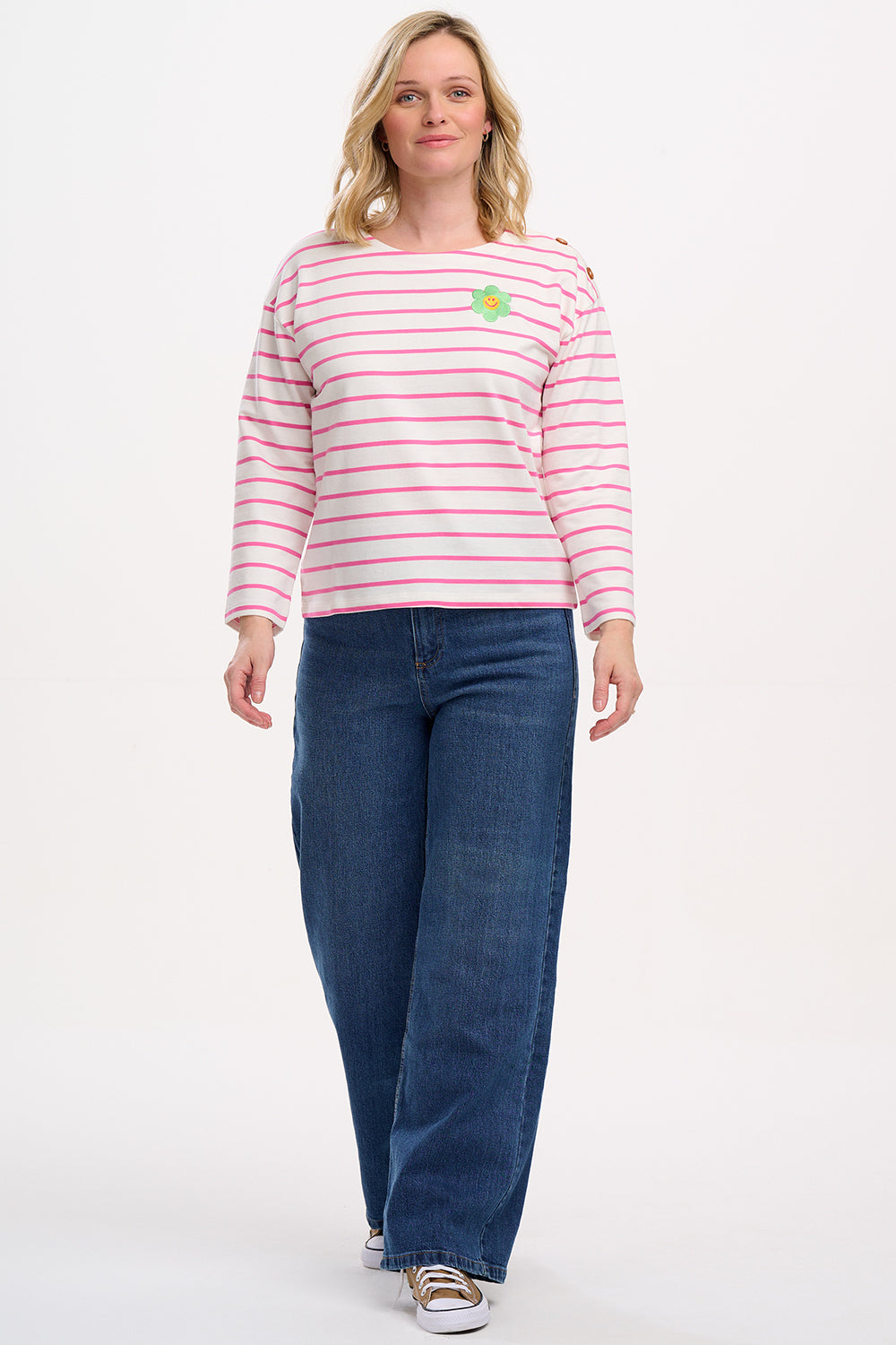A woman wearing an off-white and pink striped Breton top with a smiley flower embroidery on the chest, paired with jeans.