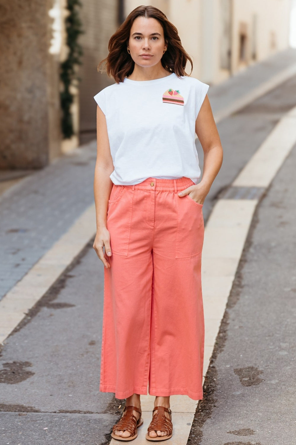Woman wearing a white t-shirt with a logo and coral pants standing on a street.