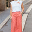 Woman wearing a white t-shirt with a logo and coral pants standing on a street.