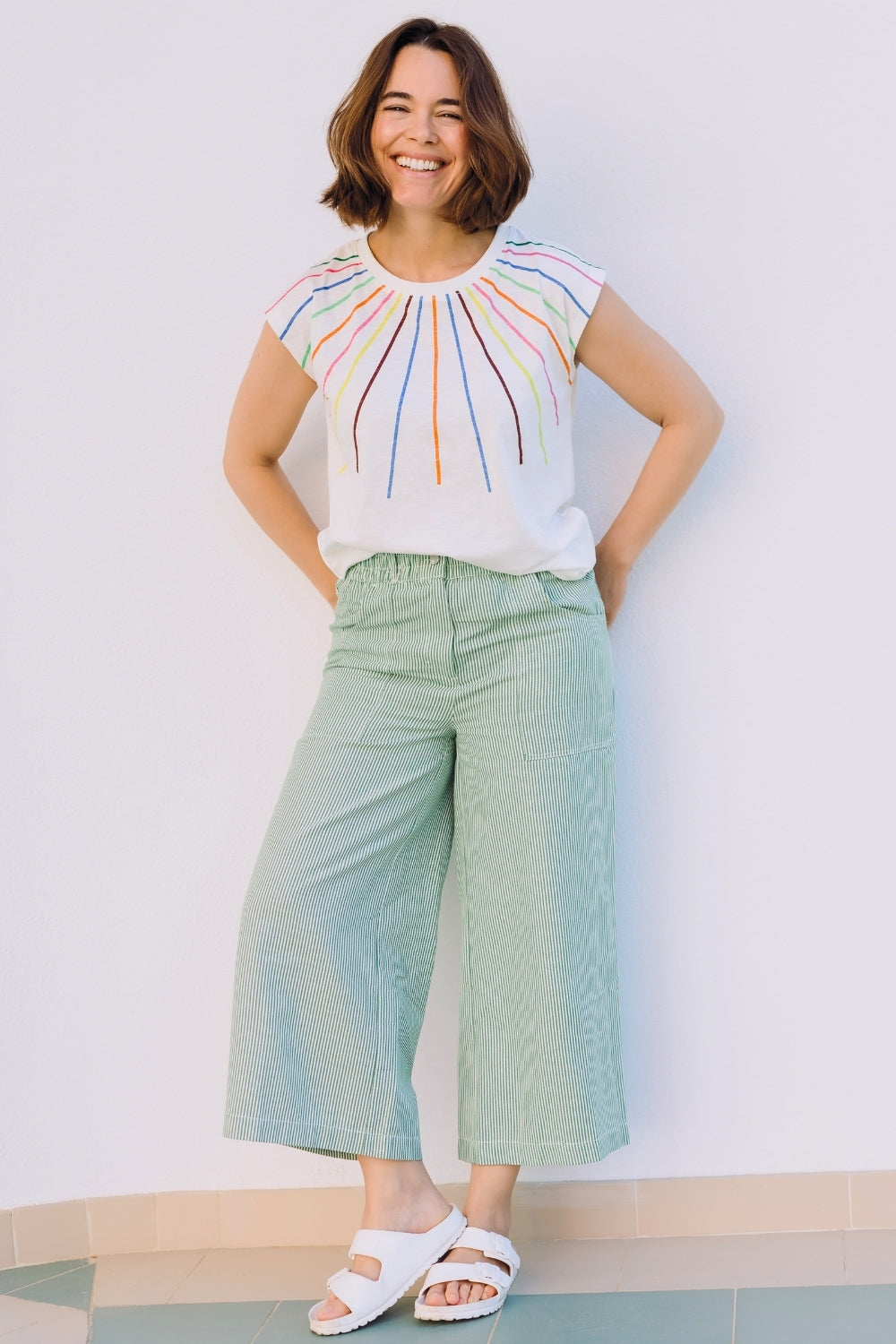 Woman wearing a colorful sleeveless top and light green pants against a plain background