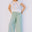 Woman wearing a colorful sleeveless top and light green pants against a plain background