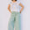 Woman wearing a colorful sleeveless top and light green pants against a plain background