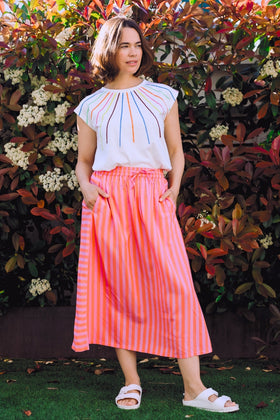 Woman wearing a colorful striped top and pink skirt standing in front of a leafy background