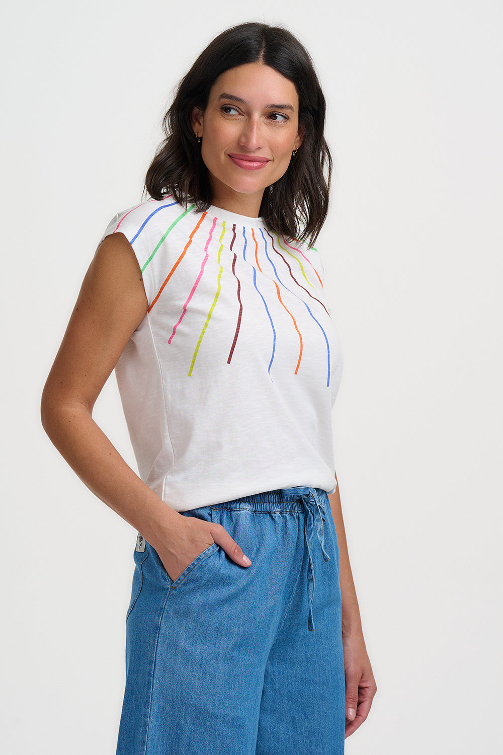 Woman wearing a white sleeveless top with colorful designs and blue pants on a white background