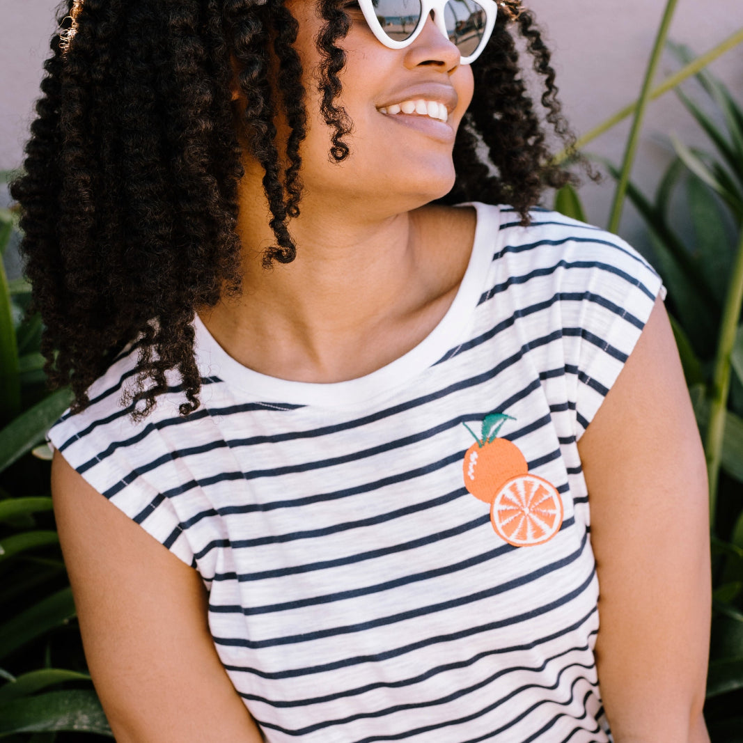 A woman smiling and wearing a sleeveless striped white and navy t-shirt with an embroidered orange on the chest.