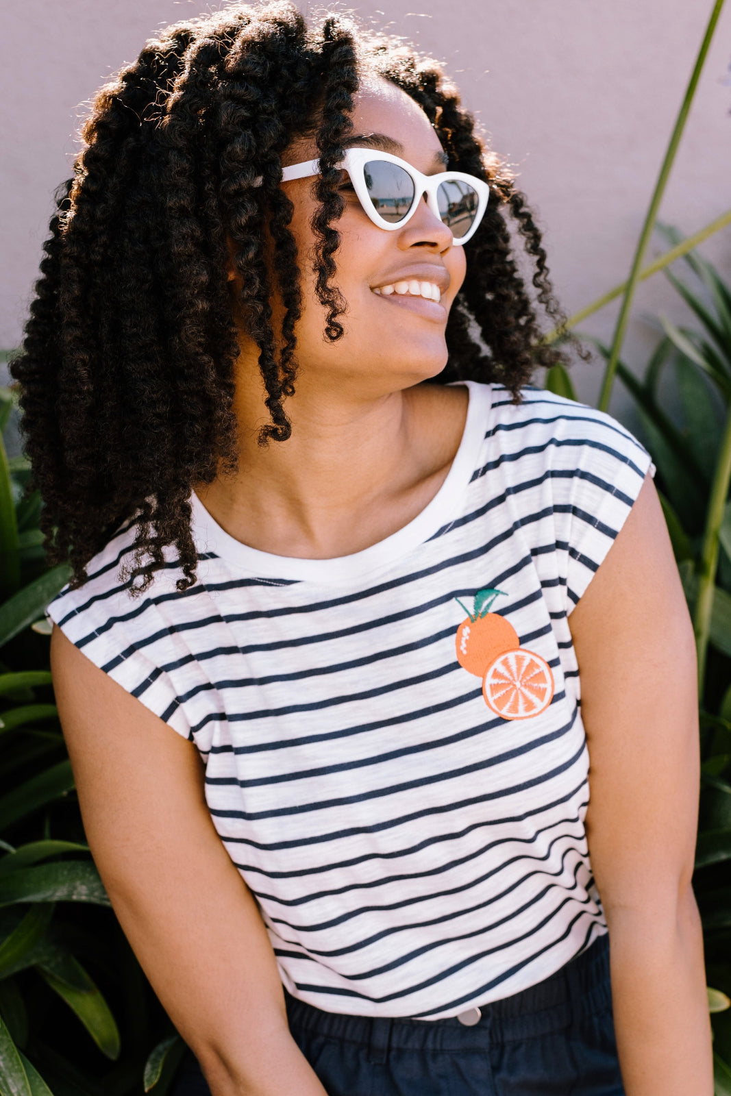 A woman smiling and wearing a sleeveless striped white and navy t-shirt with an embroidered orange on the chest.