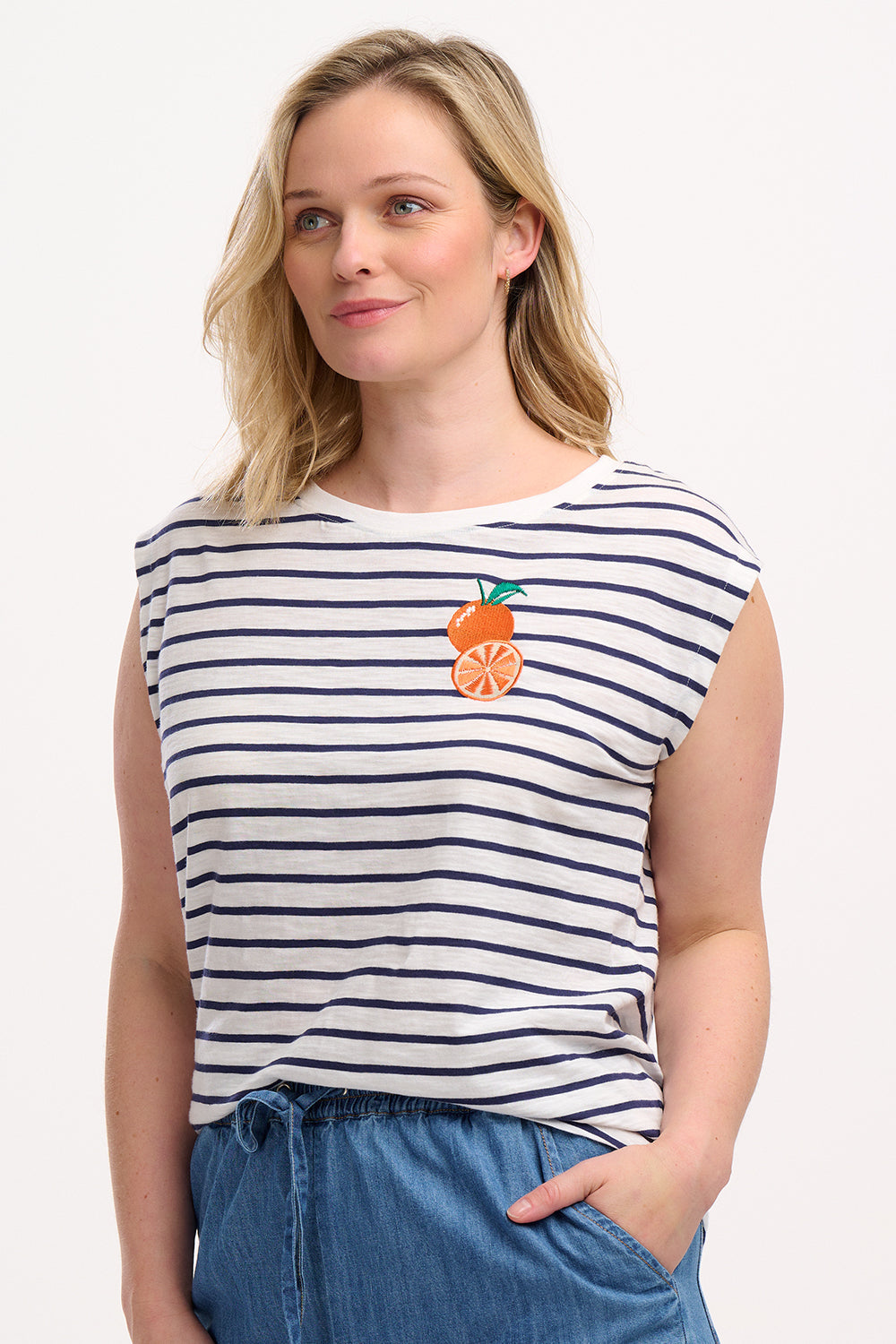 A woman wearing an off-white sleeveless tank top with navy stripes and a fruity orange embroidery on the chest.