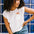 A woman wearing an off-white sleeveless t-shirt with a graphic of a sun in a pocket position, paired with blue denim jeans.