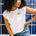A woman wearing an off-white sleeveless t-shirt with a graphic of a sun in a pocket position, paired with blue denim jeans.