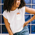 A woman wearing an off-white sleeveless t-shirt with a graphic of a sun in a pocket position, paired with blue denim jeans.