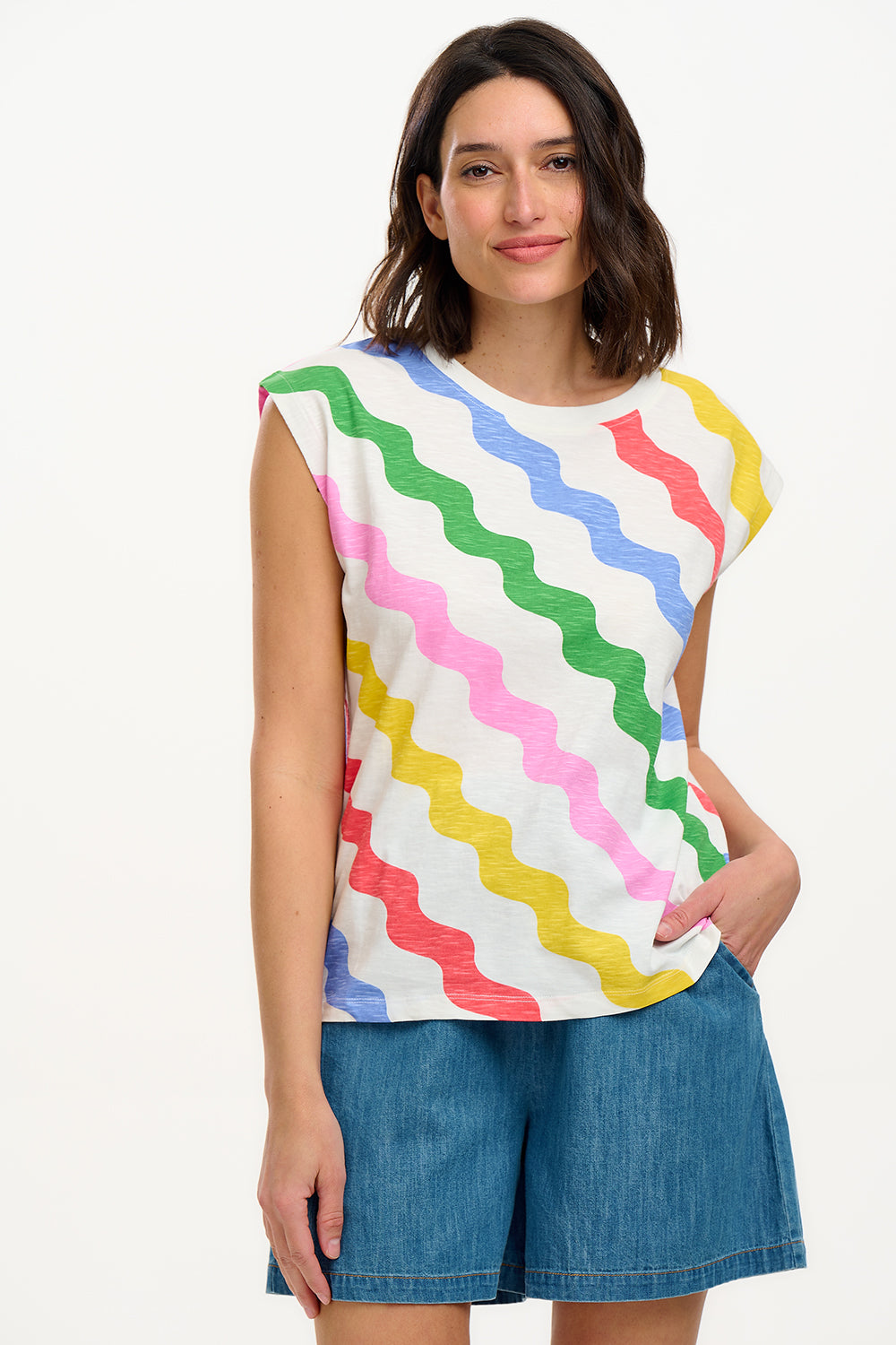 A woman modeling a sleeveless t-shirt with a geometric pattern of rainbow-colored waves on an off-white background.