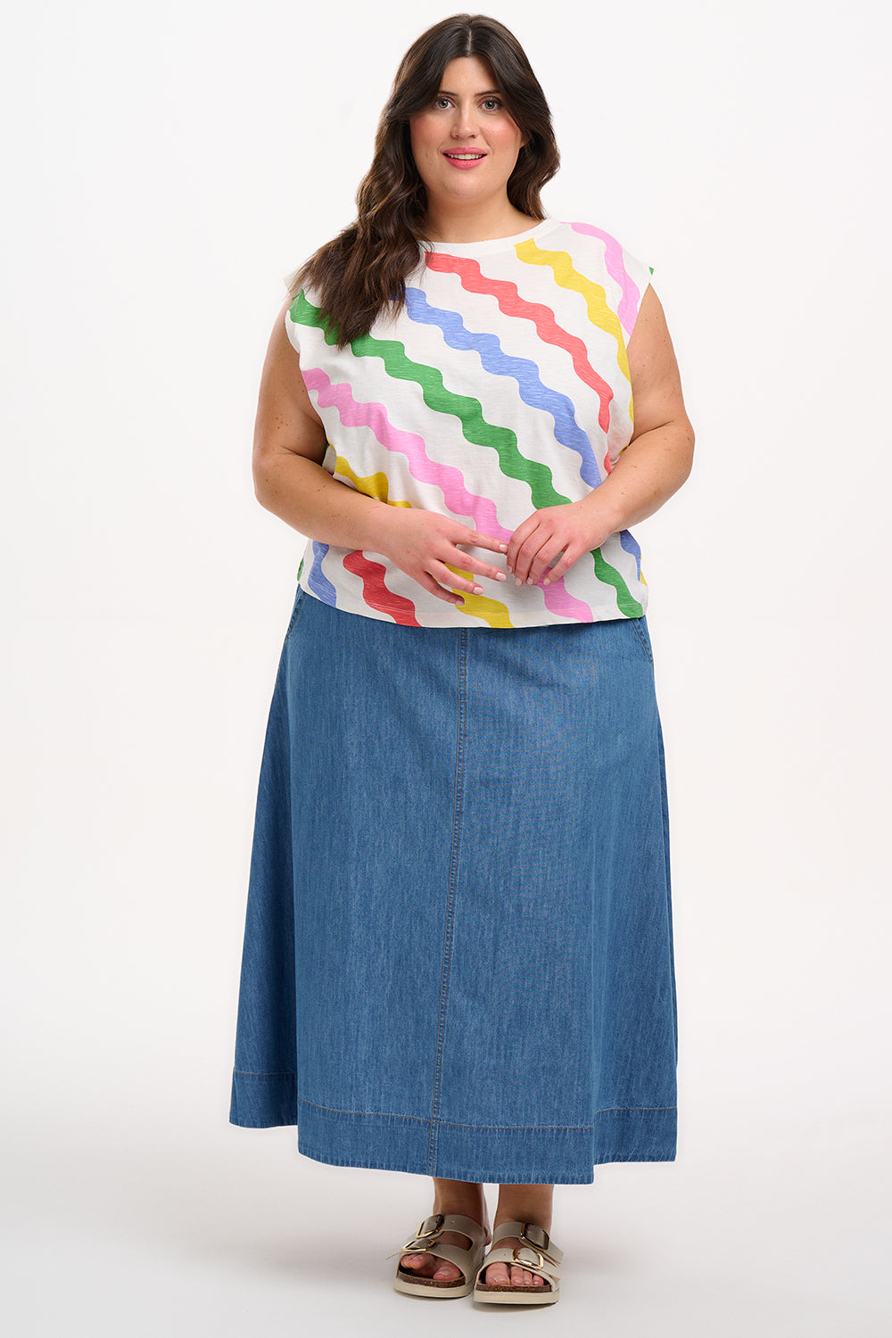 A woman modeling a sleeveless t-shirt with a geometric pattern of rainbow-colored waves on an off-white background.