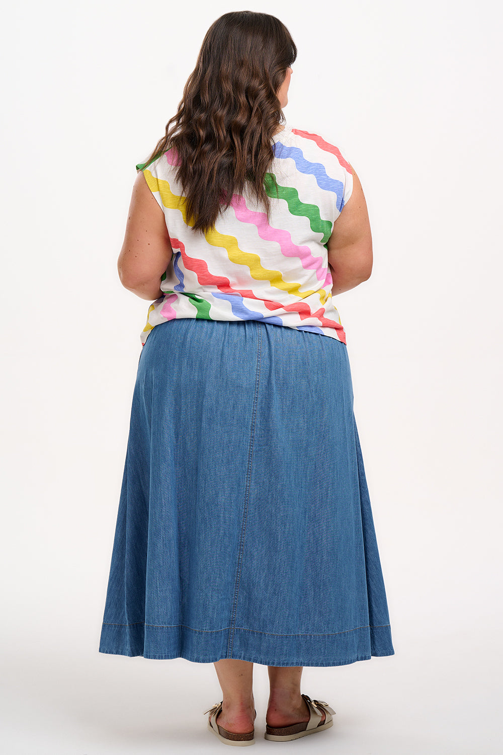 A woman modeling a sleeveless t-shirt with a geometric pattern of rainbow-colored waves on an off-white background.