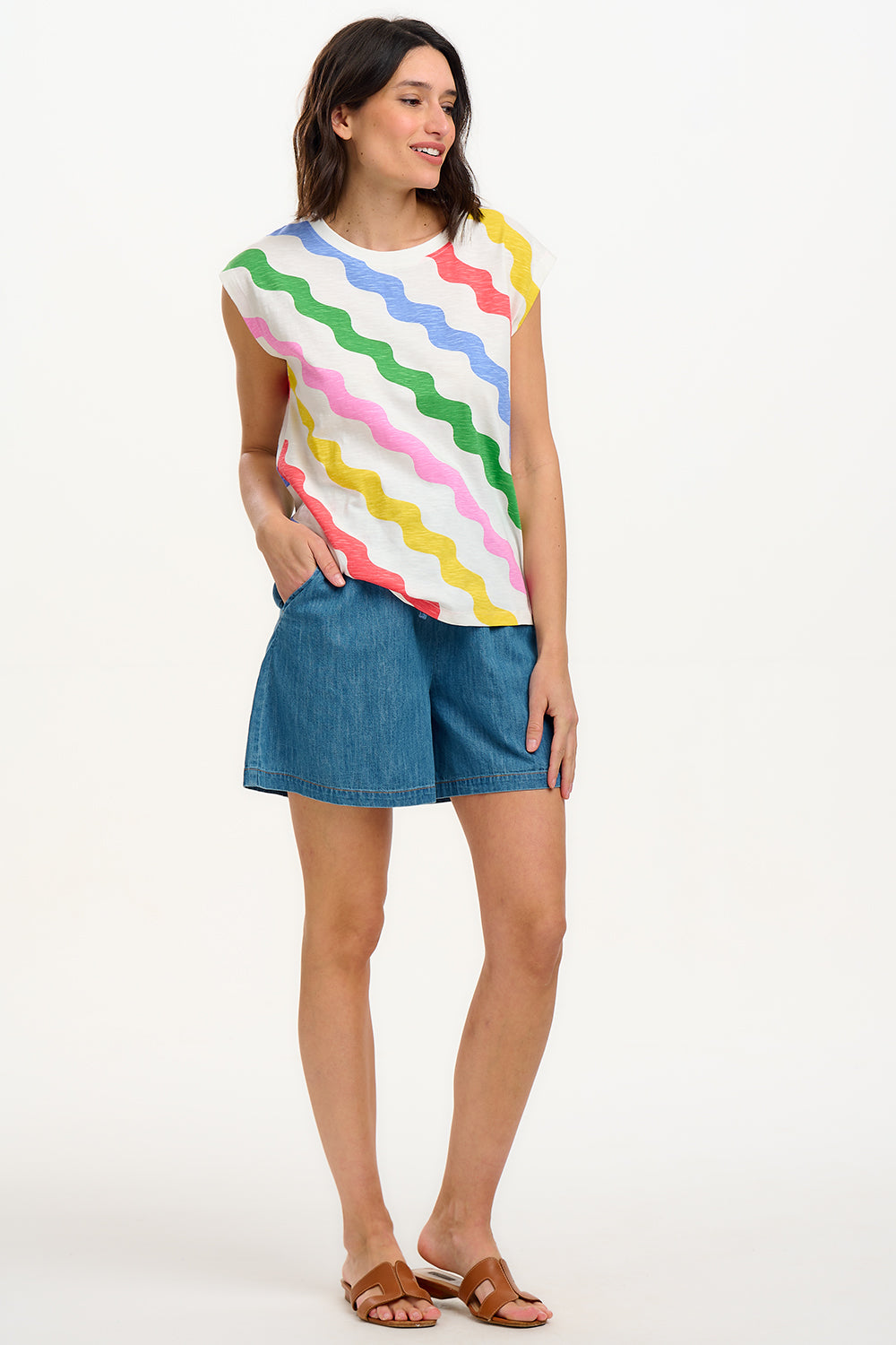 A woman modeling a sleeveless t-shirt with a geometric pattern of rainbow-colored waves on an off-white background.