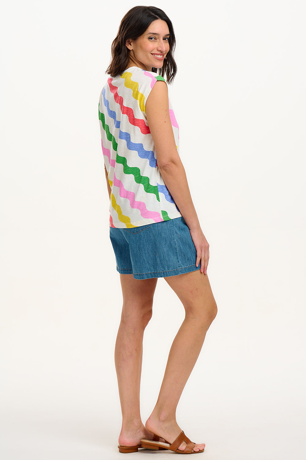 A woman modeling a sleeveless t-shirt with a geometric pattern of rainbow-colored waves on an off-white background.