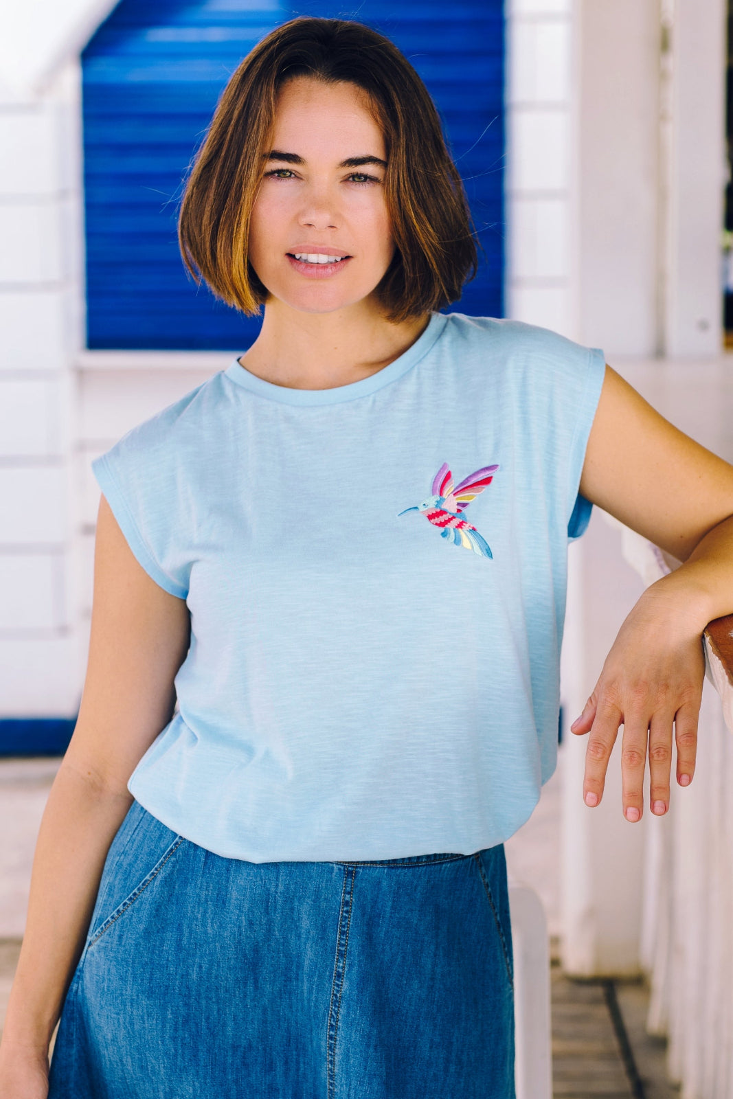 A woman wearing a pale blue sleeveless t-shirt with a tropical hummingbird graphic on the front, paired with a denim skirt.