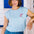 A woman wearing a pale blue sleeveless t-shirt with a tropical hummingbird graphic on the front, paired with a denim skirt.