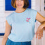 A woman wearing a pale blue sleeveless t-shirt with a tropical hummingbird graphic on the front, paired with a denim skirt.