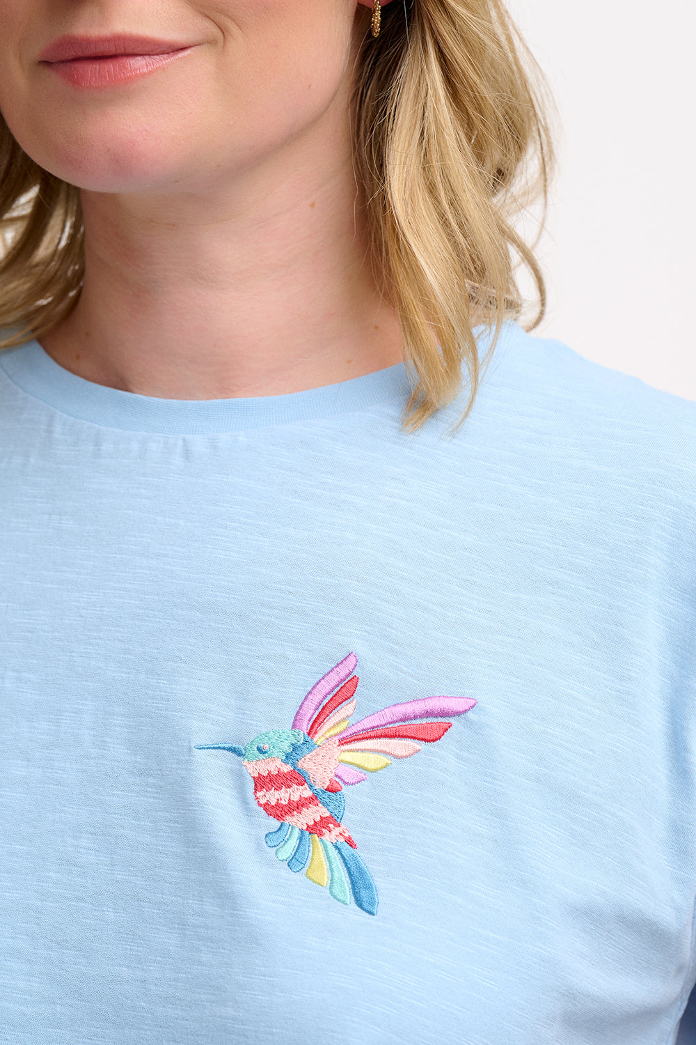 A woman wearing a pale blue sleeveless t-shirt with a tropical hummingbird graphic on the front