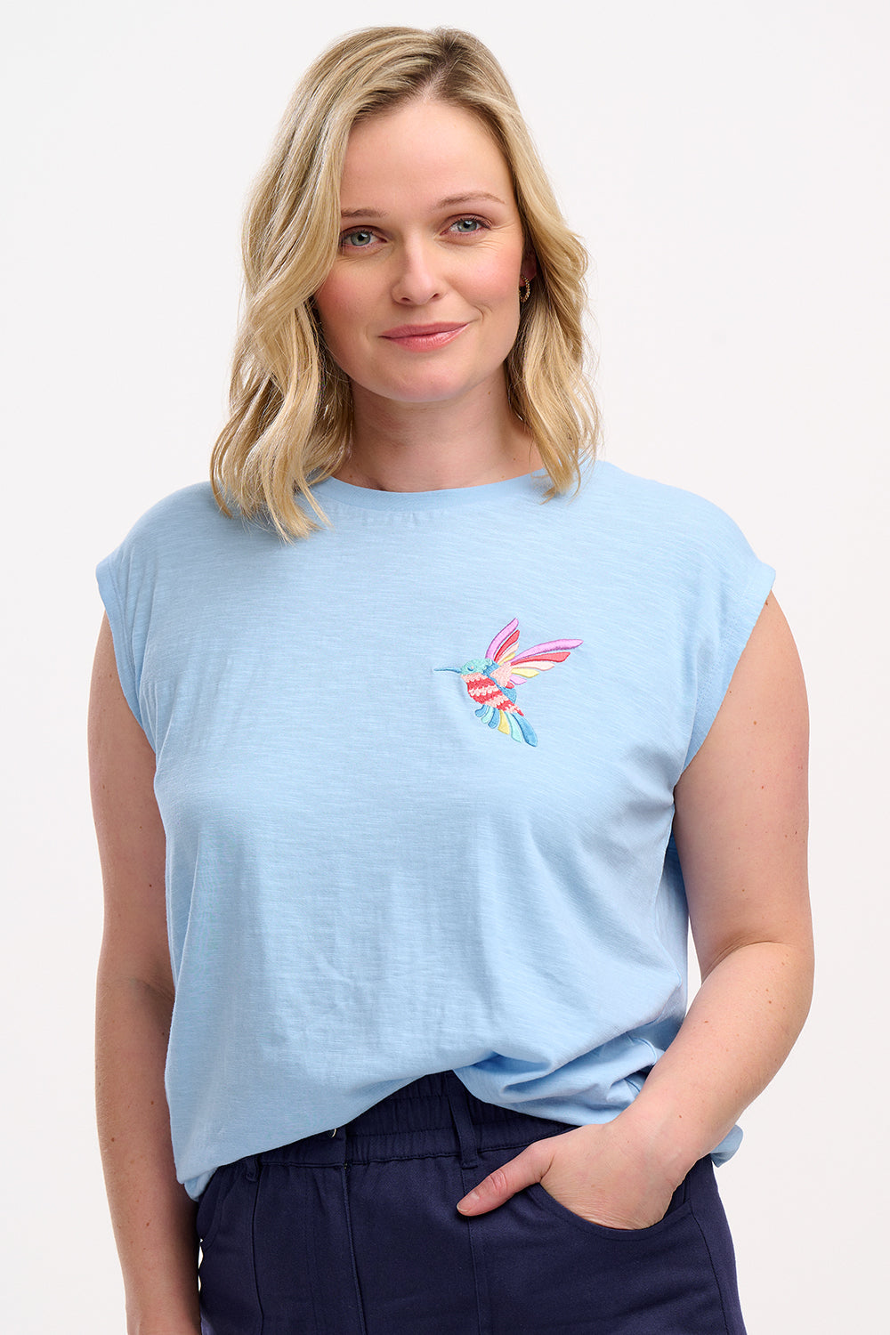 A woman wearing a pale blue sleeveless T-shirt with a tropical hummingbird graphic on the front.