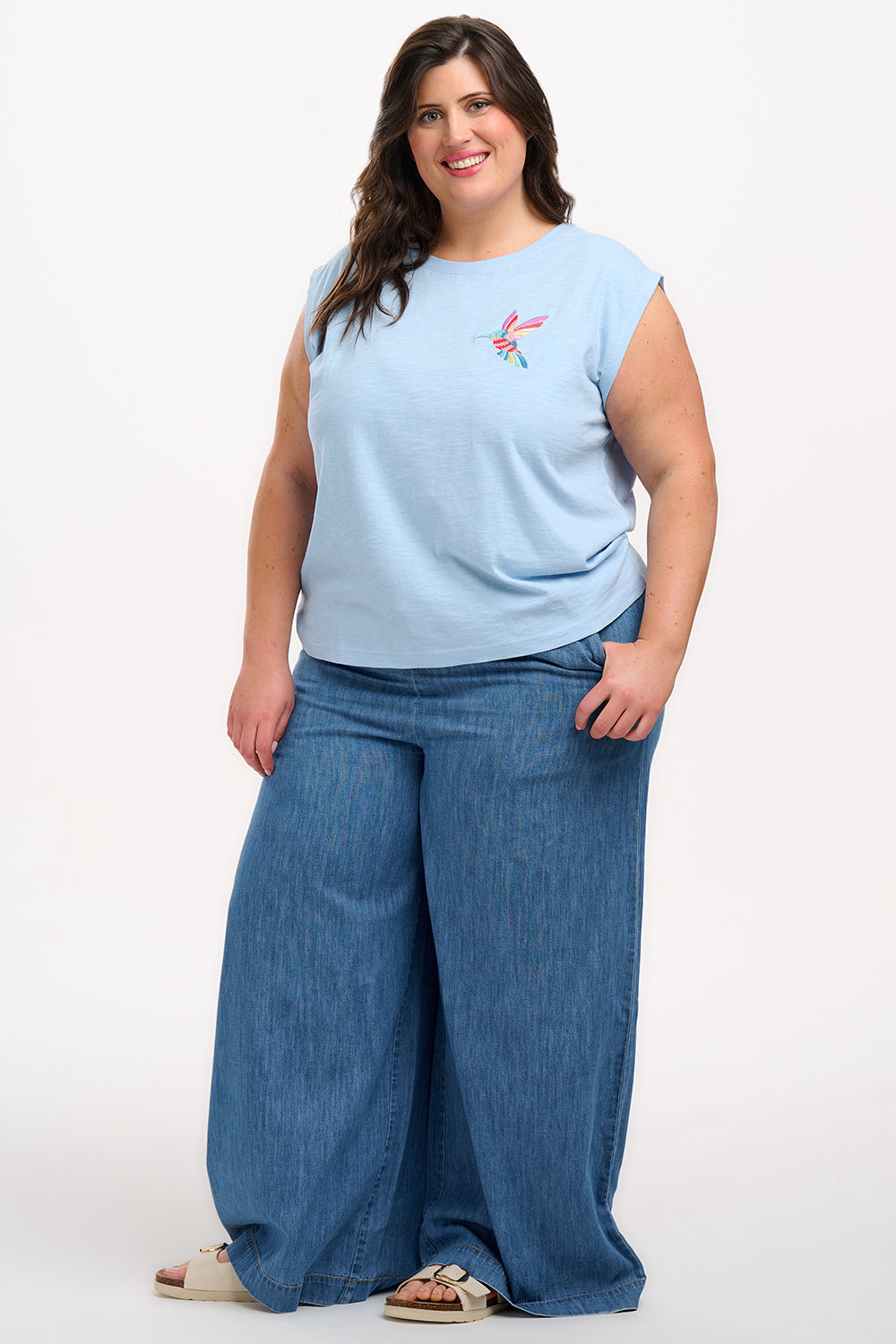 A woman wearing a pale blue sleeveless T-shirt with a tropical hummingbird graphic on the front.