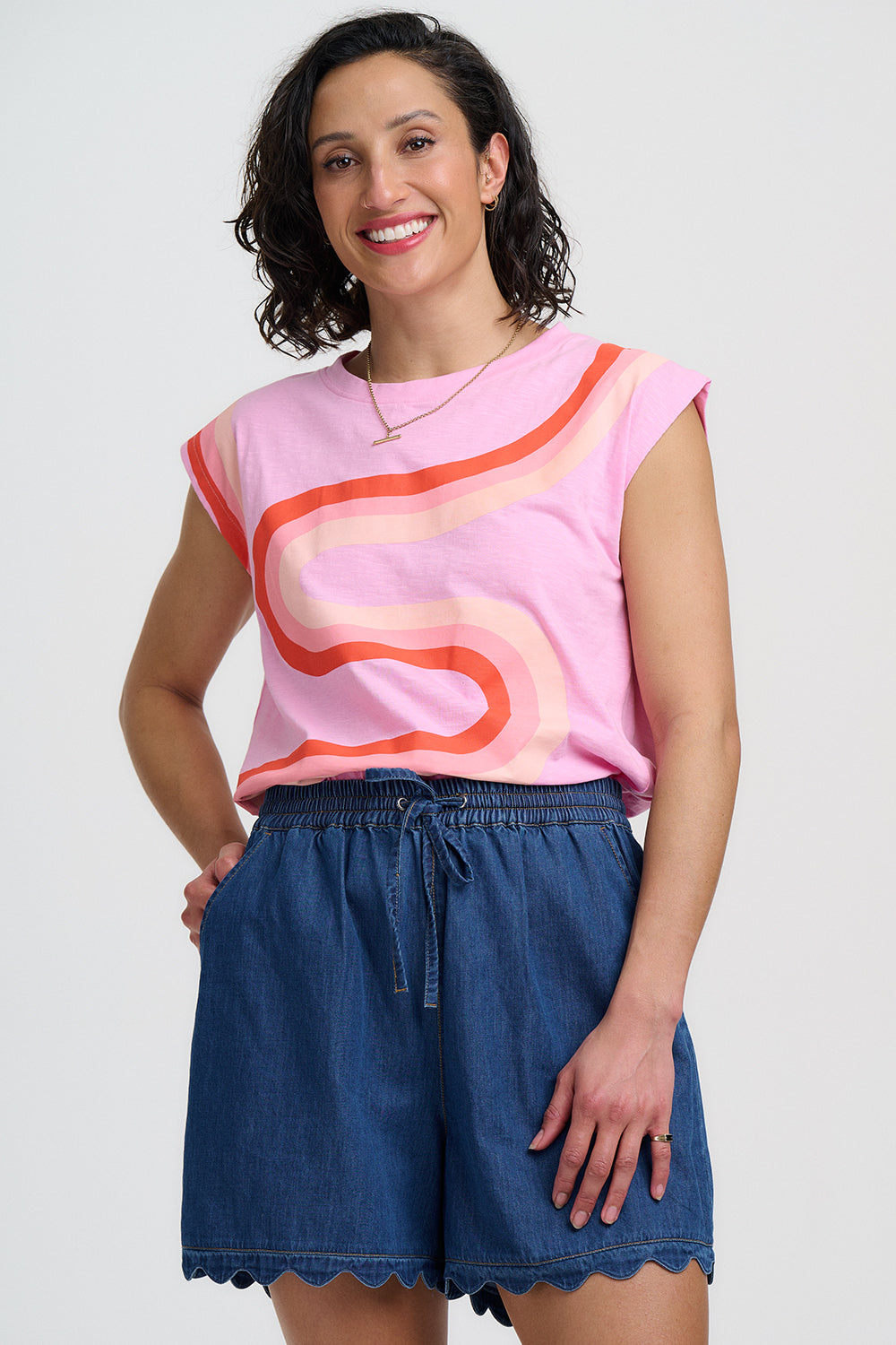 Woman wearing a pink sleeveless top with orange and blue design and blue shorts on a white background