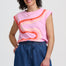 Woman wearing a pink sleeveless top with orange and blue design and blue shorts on a white background