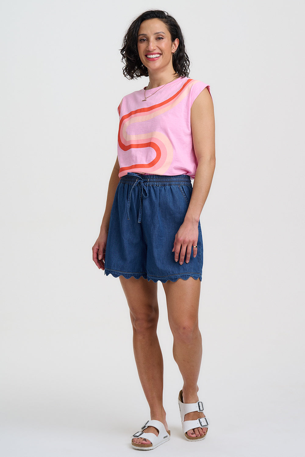 Woman wearing a pink sleeveless top with orange abstract design and blue shorts on a white background