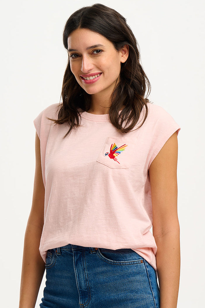 A woman smiling and wearing a pink sleeveless T-shirt with a rainbow parrot embroidery on the chest pocket, paired with blue jeans.