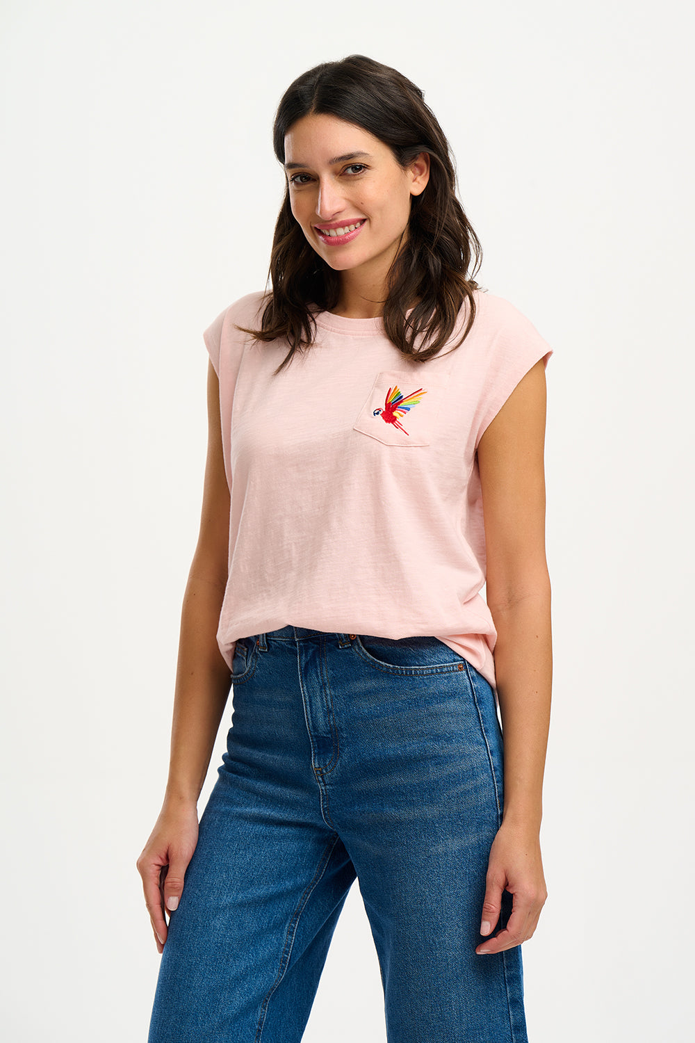 A woman smiling and wearing a pink sleeveless T-shirt with a rainbow parrot embroidery on the chest pocket, paired with blue jeans.