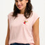 A woman smiling and wearing a pink sleeveless T-shirt with a rainbow parrot embroidery on the chest pocket, paired with blue jeans.
