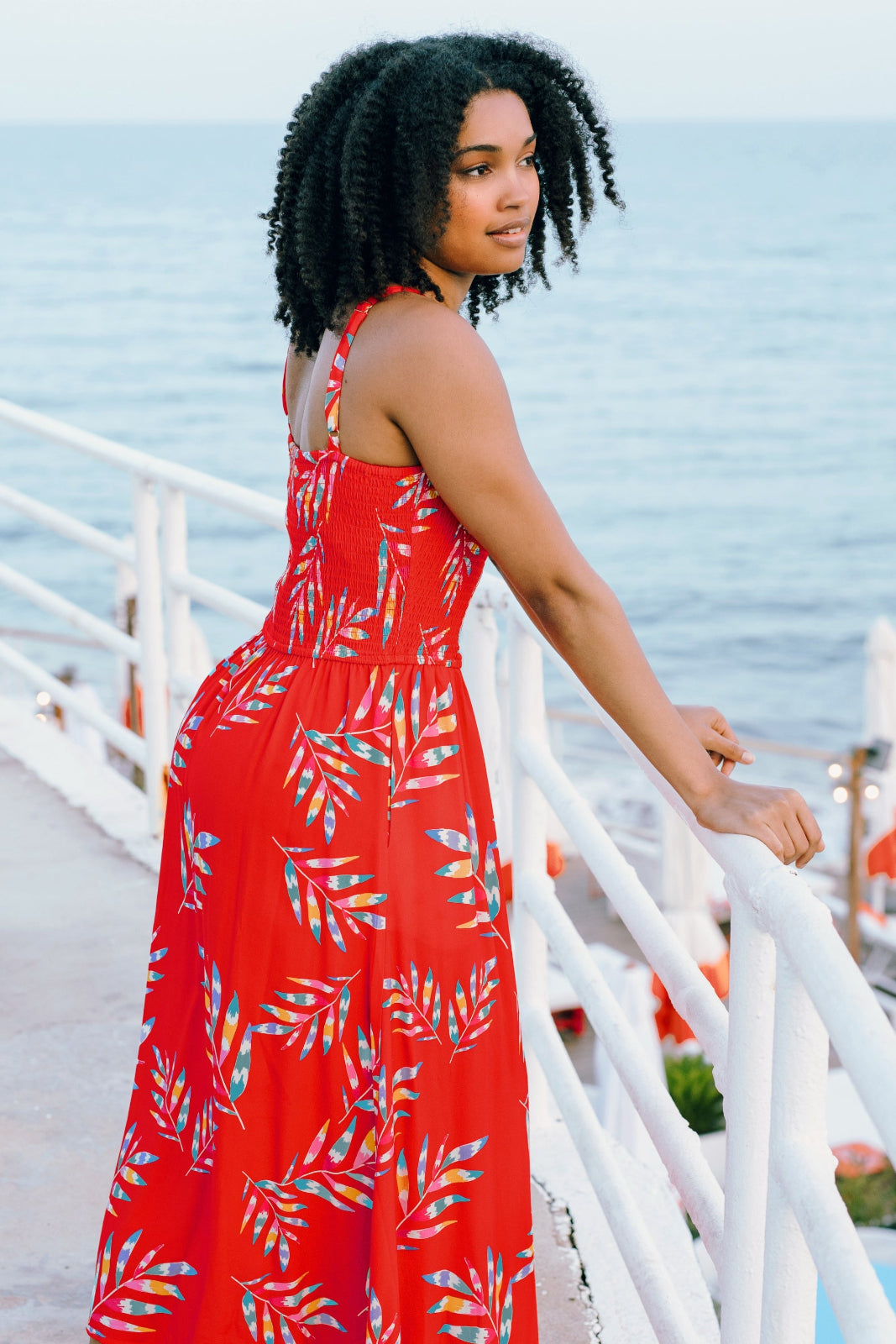 A model wearing a red midi dress with a tropical leaf print, featuring a shirred bust and pockets, standing by a coastal backdrop.