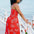 A model wearing a red midi dress with a tropical leaf print, featuring a shirred bust and pockets, standing by a coastal backdrop.
