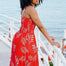 A model wearing a red midi dress with a tropical leaf print, featuring a shirred bust and pockets, standing by a coastal backdrop.