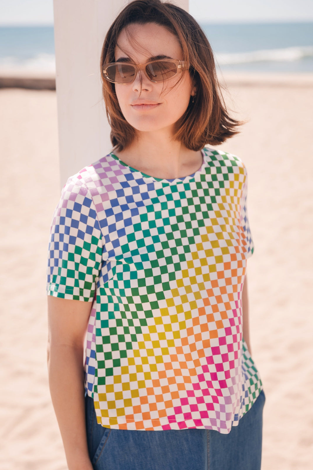 A woman standing on a beach wearing a multicolored rainbow checkerboard t-shirt with a round neckline and short sleeves.